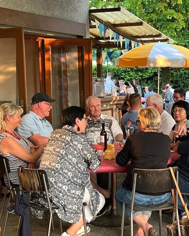 Eine Gruppe sitzt bei einem Anlass am Tisch vor dem Clublokal, im Hintergrund weitere Gäste und ein Sonnenschirm