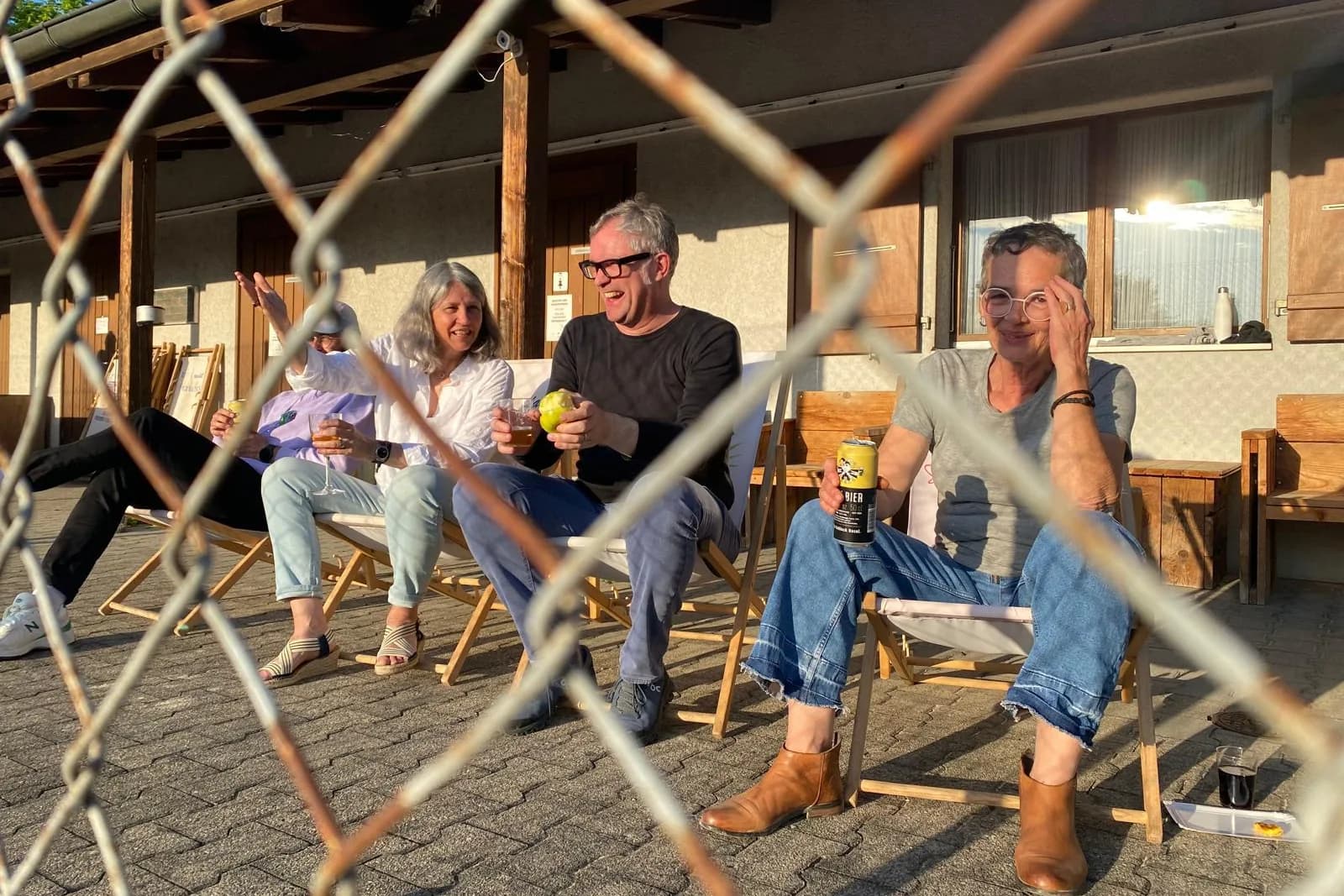 Vier Leute sitzen lachend in Liegestühlen vor dem Clublokal, Abendsonne, durch den Zaun fotografiert